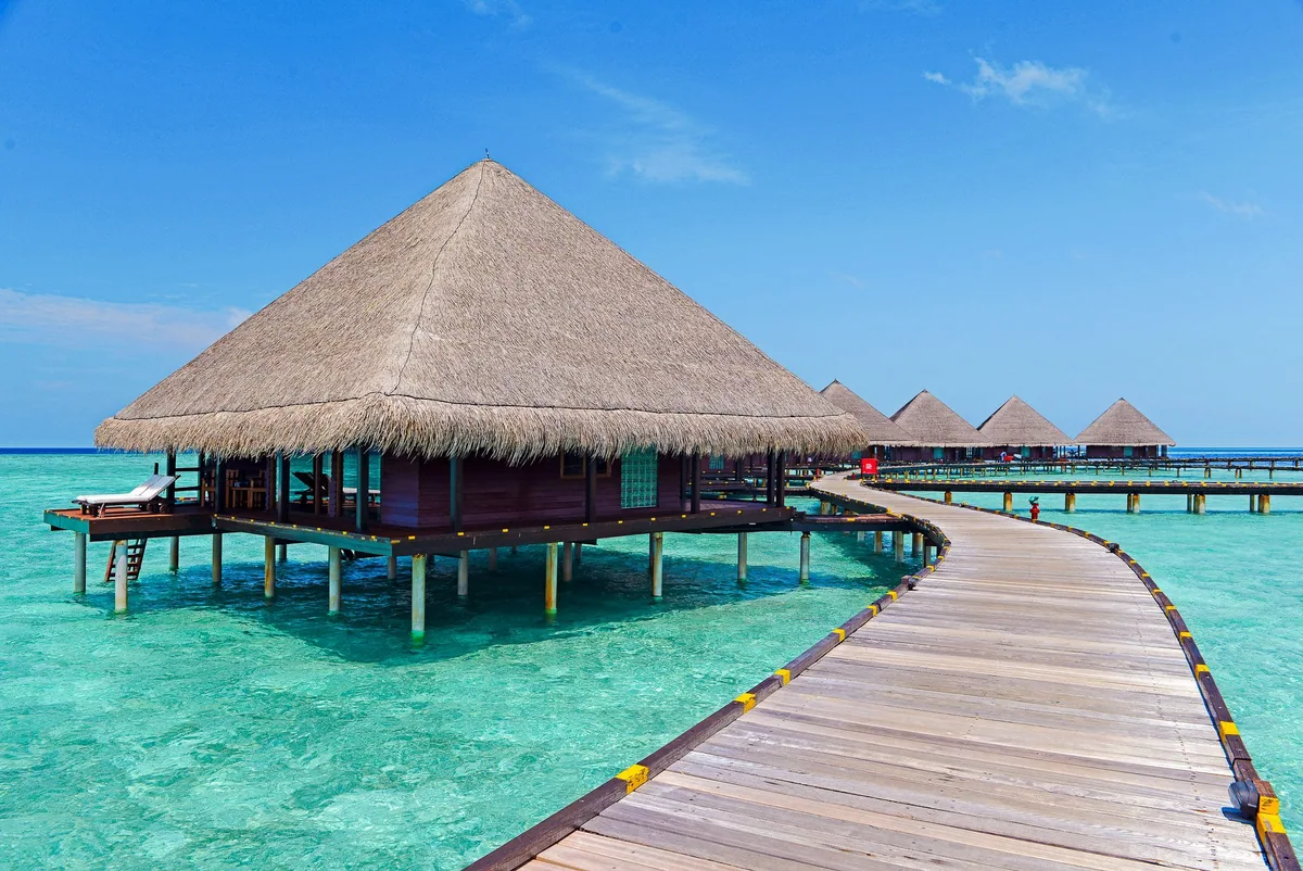 Adaaran Club Rannalhi, villa overwater, South Malé Atoll, Maldives, Water Villa row exterior at midday, the large pyramidal thatched-roof villa on stilts (the property's signature pyramid Water Villa geometry, distinct from the more common pitched-roof villas nearby), the long timber boardwalk tracking past the row of villas, and the crystal turquoise lagoon below.