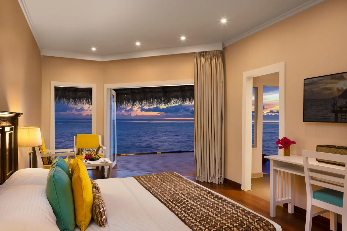 Adaaran Club Rannalhi, villa overwater, South Malé Atoll, Maldives, Water Villa interior at twilight, the cream-painted walls with recessed ceiling lights, a yellow accent armchair beside the open glazed door panels framing the open ocean horizon, multi-coloured throw cushions on the king bed (green, yellow, patterned), a side dressing desk with framed-photo wall art, and warm bedside lamp glow.