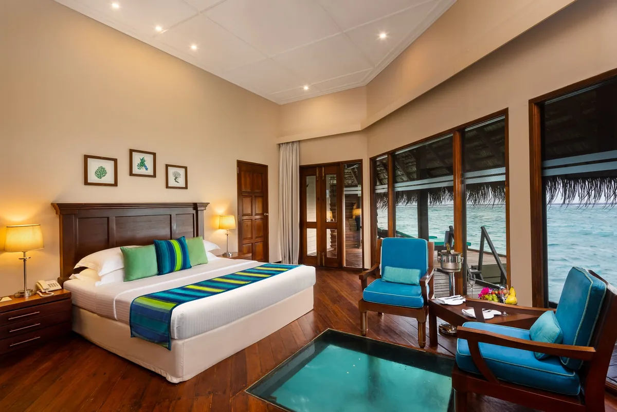 Adaaran Club Rannalhi, villa overwater, South Malé Atoll, Maldives, Sunset Water Villa interior, the dark-hardwood floor with inset GLASS-FLOOR PANEL revealing the lagoon below (the signature transparent-floor feature), royal-blue armchair pair at the corner glazing opening onto neighbouring villas, the wood-framed picture wall above the bed, and a blue-and-green striped throw across the king linens.