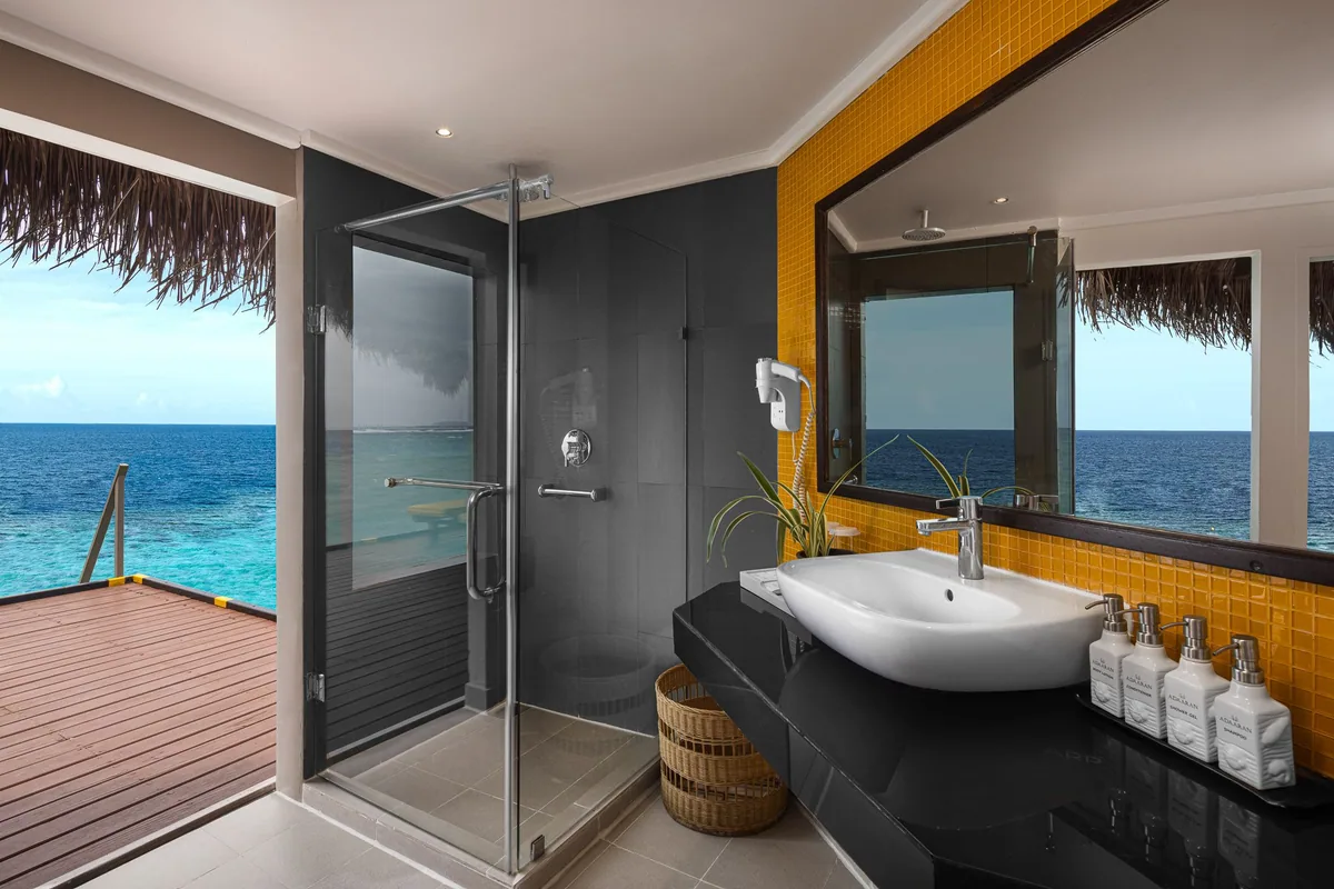 Adaaran Club Rannalhi, villa overwater, South Malé Atoll, Maldives, Water Villa bathroom, the yellow-mosaic feature wall behind the framed vanity mirror, dark-stone counter with twin vessel sink, the floor-to-ceiling glass-walled rain shower with the reef view through the open door, and the timber sundeck beyond.