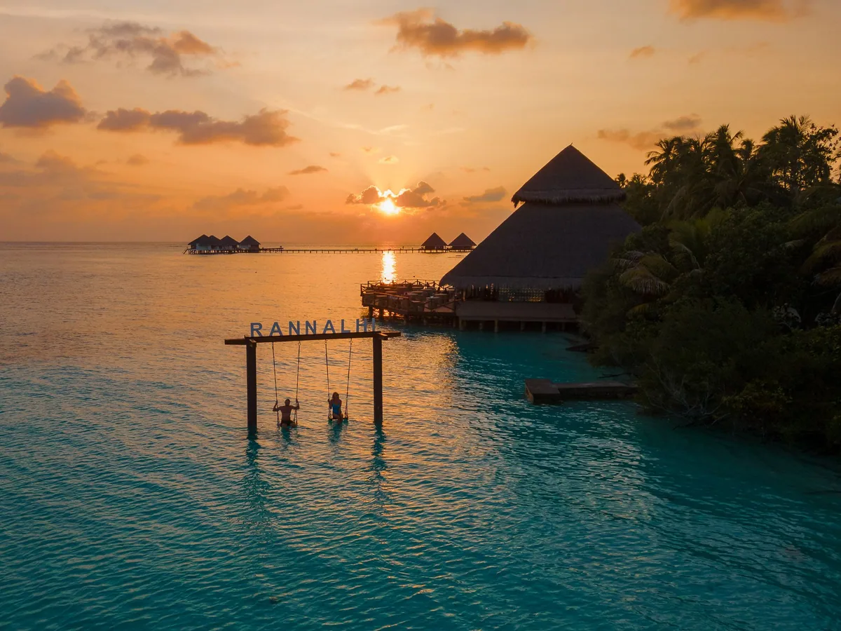 Adaaran Club Rannalhi, lifestyle, South Malé Atoll, Maldives, The RANNALHI wordmark beach swing at sunset, the freestanding wooden swing-frame in the lagoon shallows with two timber plank seats just above the water, a couple mid-swing in silhouette, the over-water dining pavilion at the right, the jetty extending to the distant over-water villa row, and the deep orange sun on the horizon.