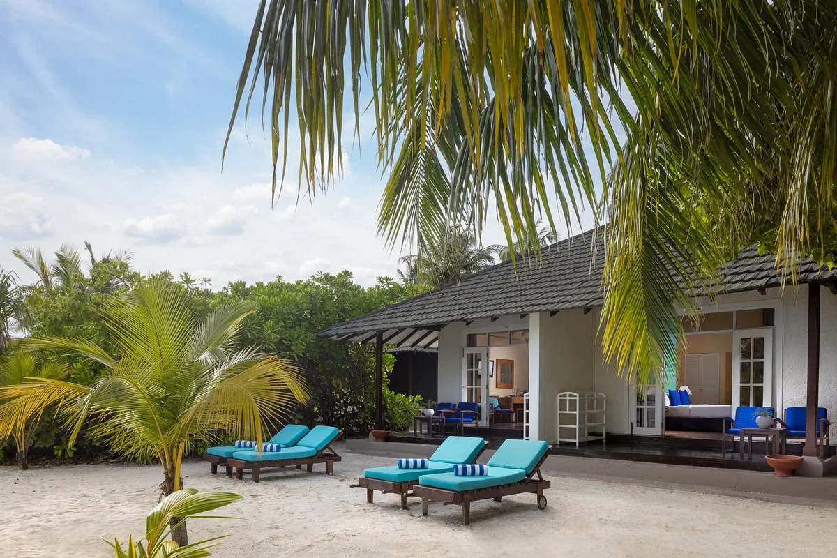 Adaaran Meedhupparu, Two Bedroom Family Beach Villa with dark-tile sloped roof, white walls, covered verandah with blue armchairs and sun loungers on sand, Raa Atoll, Maldives