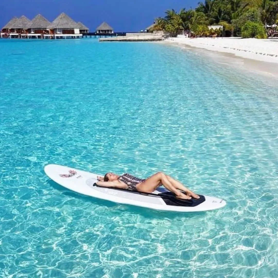 Adaaran Meedhupparu, stand-up paddleboarding on shallow lagoon with Prestige Water Villa thatched-pyramid cluster visible across lagoon, Raa Atoll, Maldives