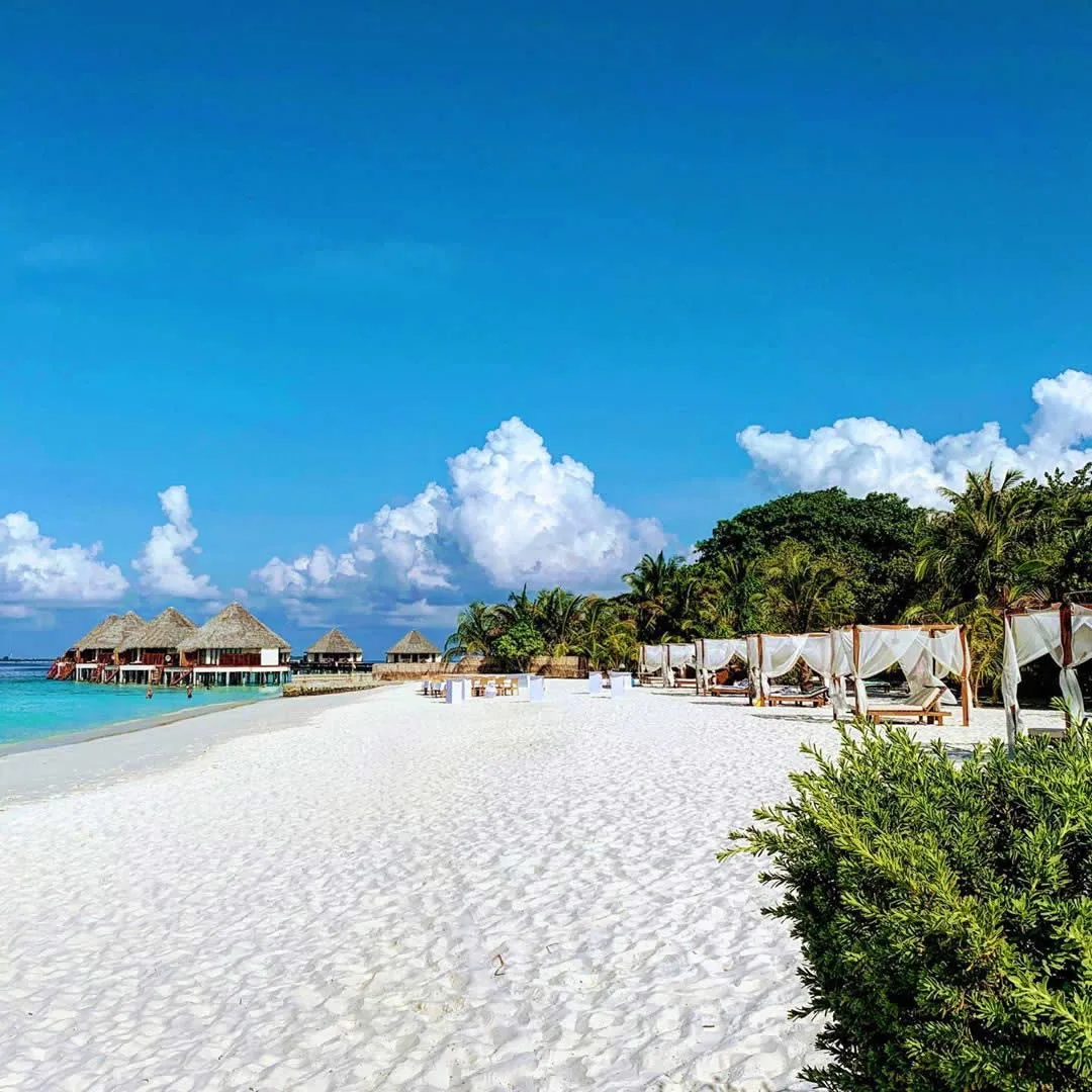 Adaaran Meedhupparu, white-sand beach spit with curtained four-poster daybeds and beach loungers, Prestige Water Villa cluster visible across lagoon, Raa Atoll, Maldives