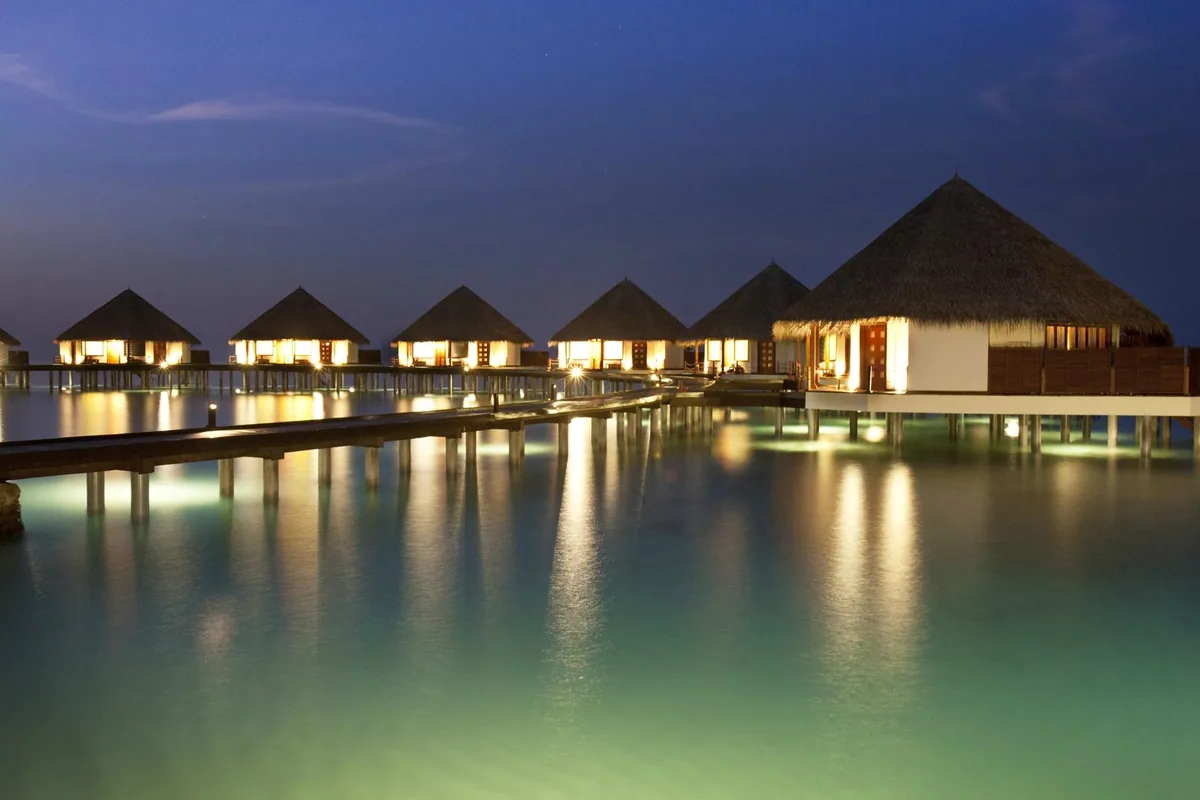 Adaaran Prestige Water Villas at twilight, row of lit thatched-pyramid villas on stilts with reflections on glass-calm lagoon, Raa Atoll, Maldives
