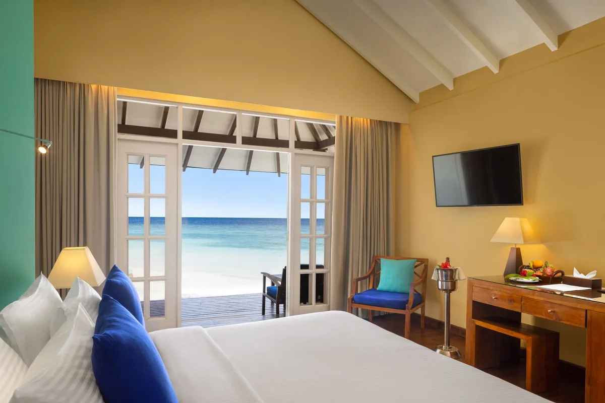 Adaaran Select Beach Villa interior with yellow walls, teal accent wall, vaulted white ceiling, king bed and french doors to beach and ocean, Raa Atoll, Maldives