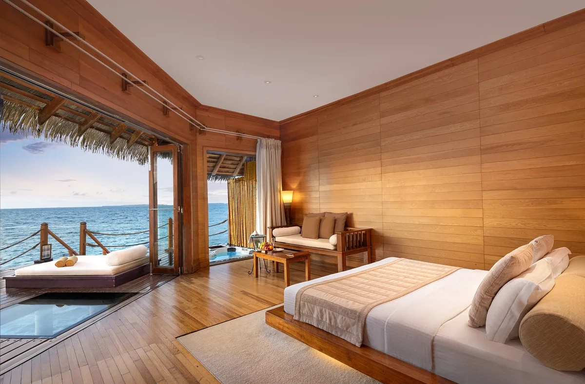 Adaaran Prestige Sunset Water Villa interior with timber wall paneling, glass-floor panel, low-platform king bed and daybed banquette with thatched-eave terrace, Raa Atoll, Maldives
