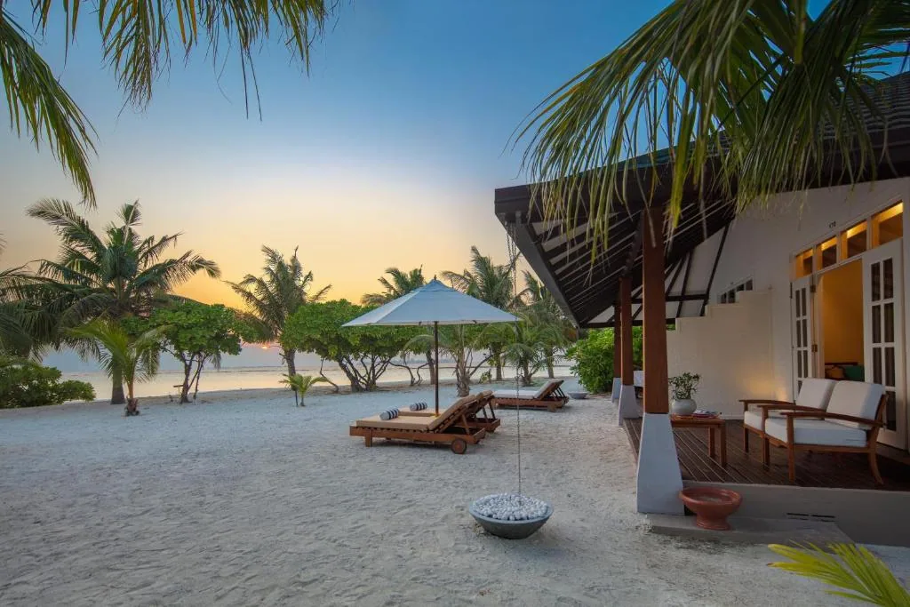 Adaaran Select Beach Villa at twilight, beachfront wooden deck with rattan sofa, sun loungers under white parasol, white-stone firepit and palms against sunset, Raa Atoll, Maldives