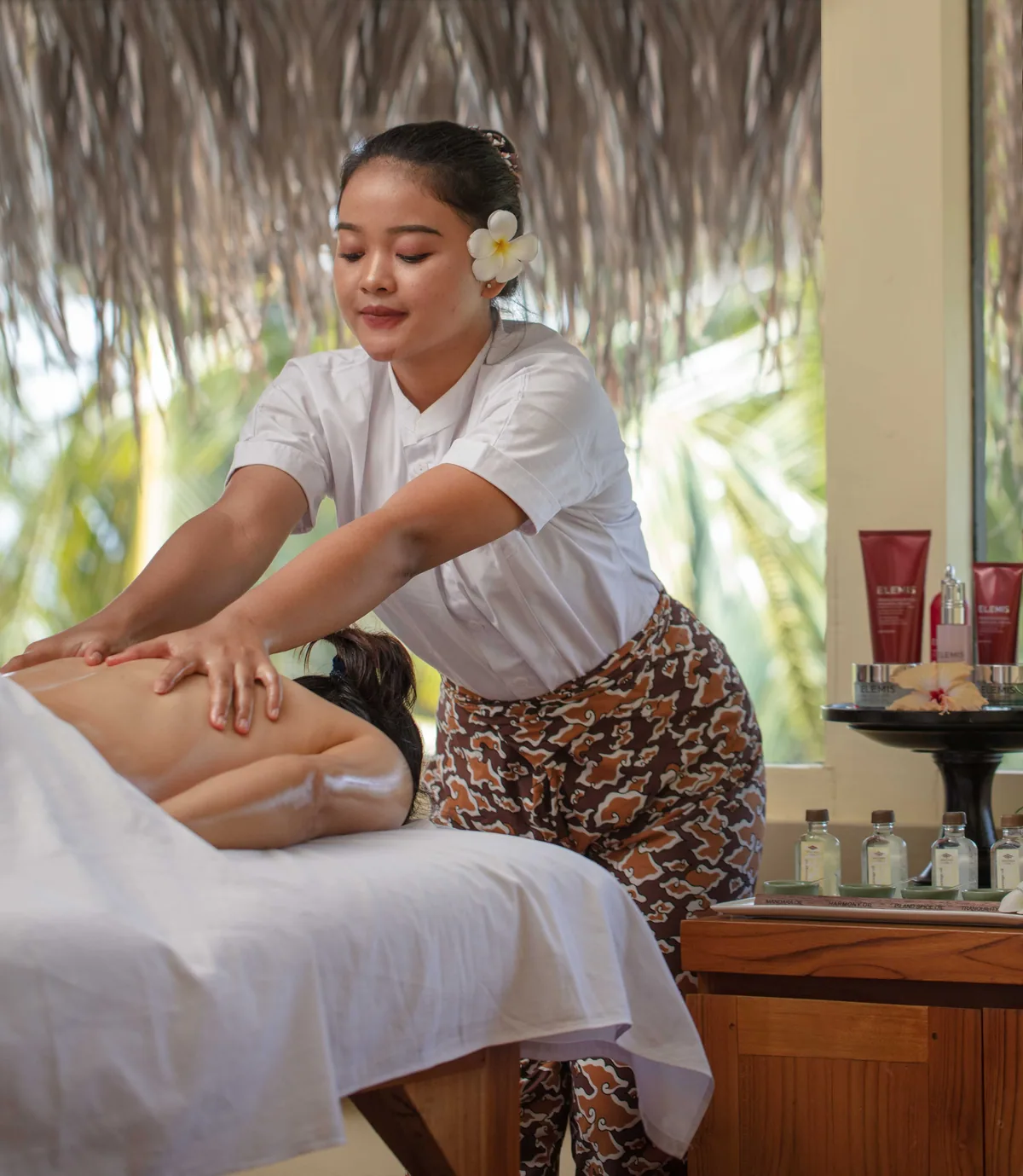Adaaran Prestige Vadoo, spa, South Malé Atoll, Maldives, Mandara Spa treatment, the spa therapist (frangipani in hair) performing the signature back massage on a guest face-down on the white-linen massage table, the row of Mandara-branded oil bottles on the timber side table, fresh frangipani arrangements, and the thatched-pavilion roof structure above.