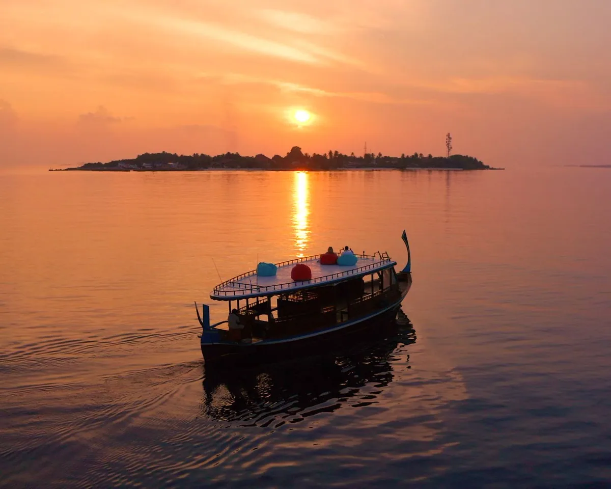Adaaran Prestige Vadoo, lifestyle, South Malé Atoll, Maldives, Sunset cruise excursion, the traditional Maldivian dhoni vessel silhouetted against a deep-orange sunset, two guests on the roof loungers in red and blue bean bags, the distant inhabited island in the background, and the still ocean reflecting the dusk colour.