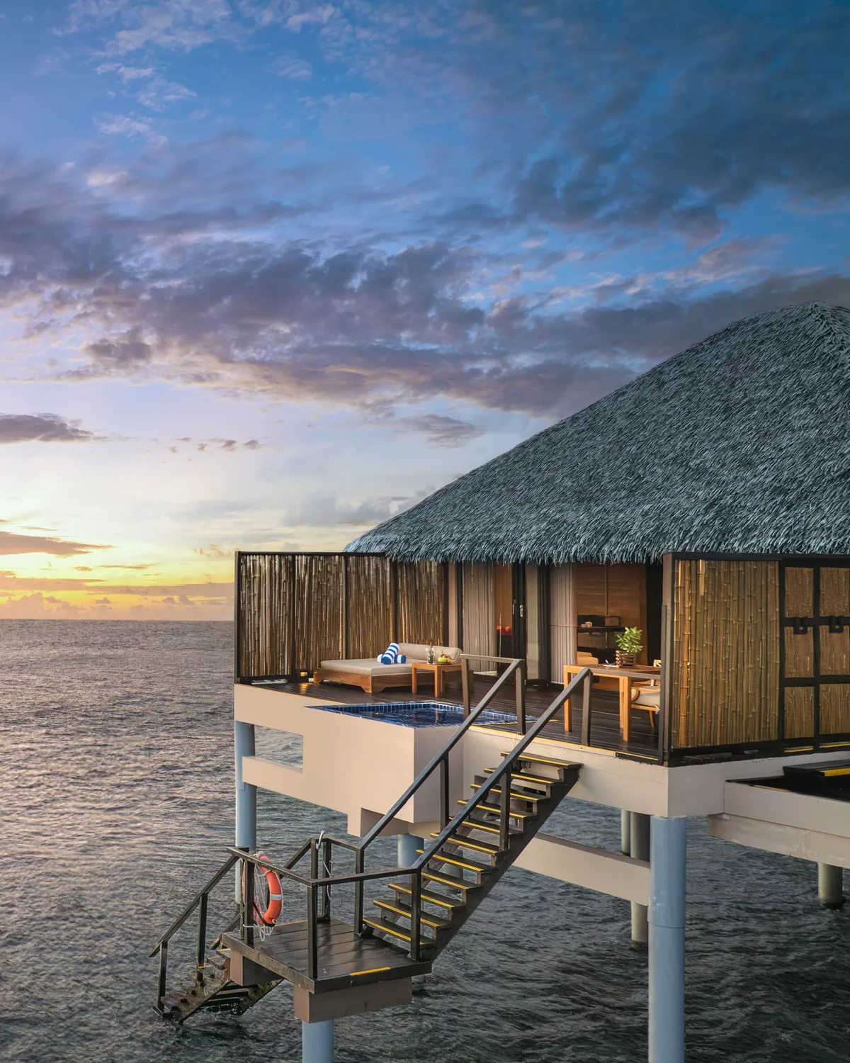 Adaaran Prestige Vadoo, villa overwater, South Malé Atoll, Maldives, Sunset Water Villa exterior at twilight, the standalone thatched-pitched-roof villa with bamboo screening, the corner glass-tile plunge pool at the deck edge, the open-air stairs dropping directly to the ocean access, and the pink-blue sunset sky behind.