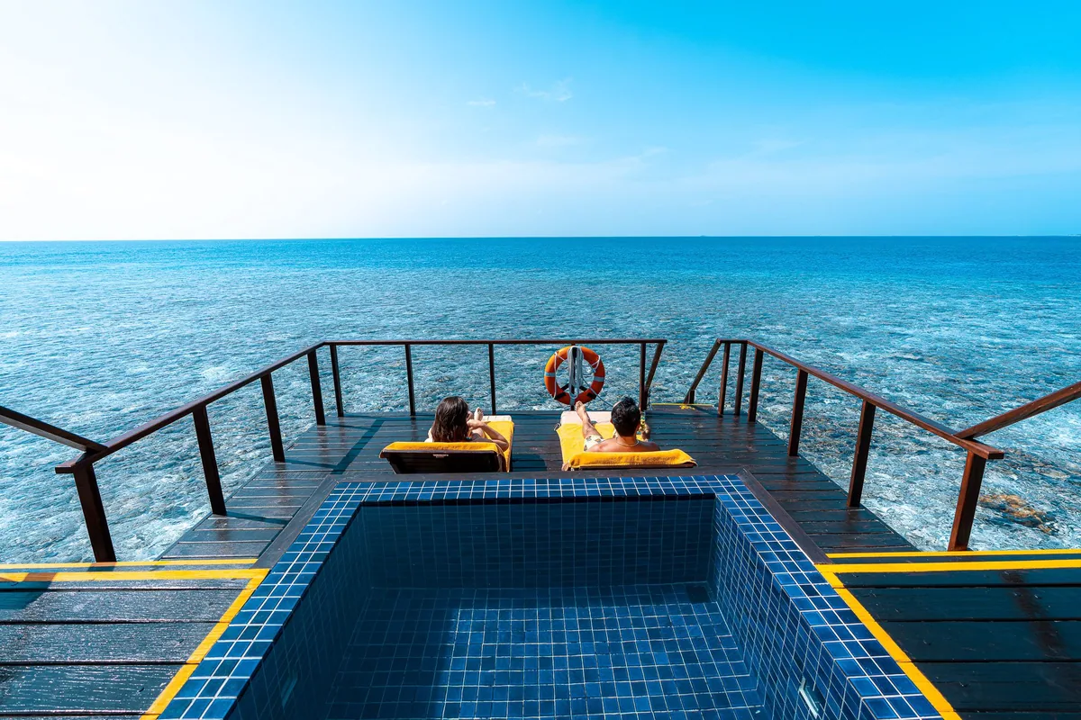 Adaaran Prestige Vadoo, villa overwater, South Malé Atoll, Maldives, Honeymoon Villa pool deck top-down view, the deep blue mosaic-tiled plunge pool extending to the deck edge, a couple on yellow-cushioned loungers facing the reef-edge ocean, a red life ring buoy on the railing, and the timber deck framing the composition.