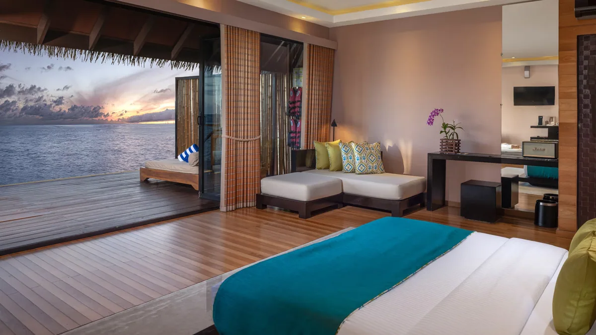 Adaaran Prestige Vadoo, villa overwater, South Malé Atoll, Maldives, Over-Water Villa with Jacuzzi bedroom interior, the pitched timber-rafter ceiling, the retractable wall opening directly to the deck and sunset ocean, Maldivian-pattern accent cushions on the modular bench, the teal throw across the king bed, an orchid in a low vase, and the warm hardwood floor.