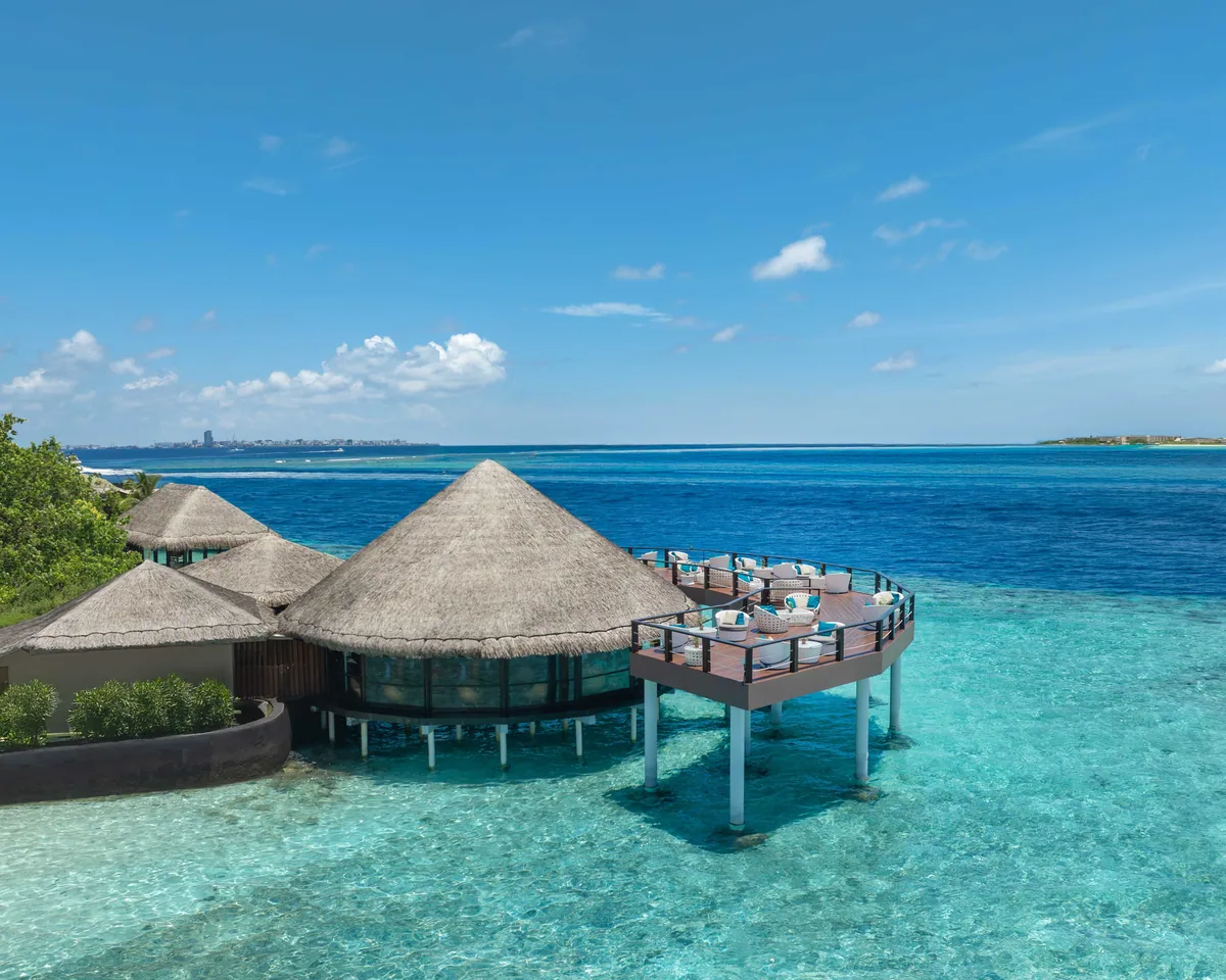 Adaaran Prestige Vadoo, dining, South Malé Atoll, Maldives, Kandholi Sky Bar over-water configuration, three thatched cone-roof buildings on stilts with an extended timber deck, lounge seating with white cushions facing the lagoon, the Malé skyline visible across the channel, and the crystal turquoise reef shallows below.