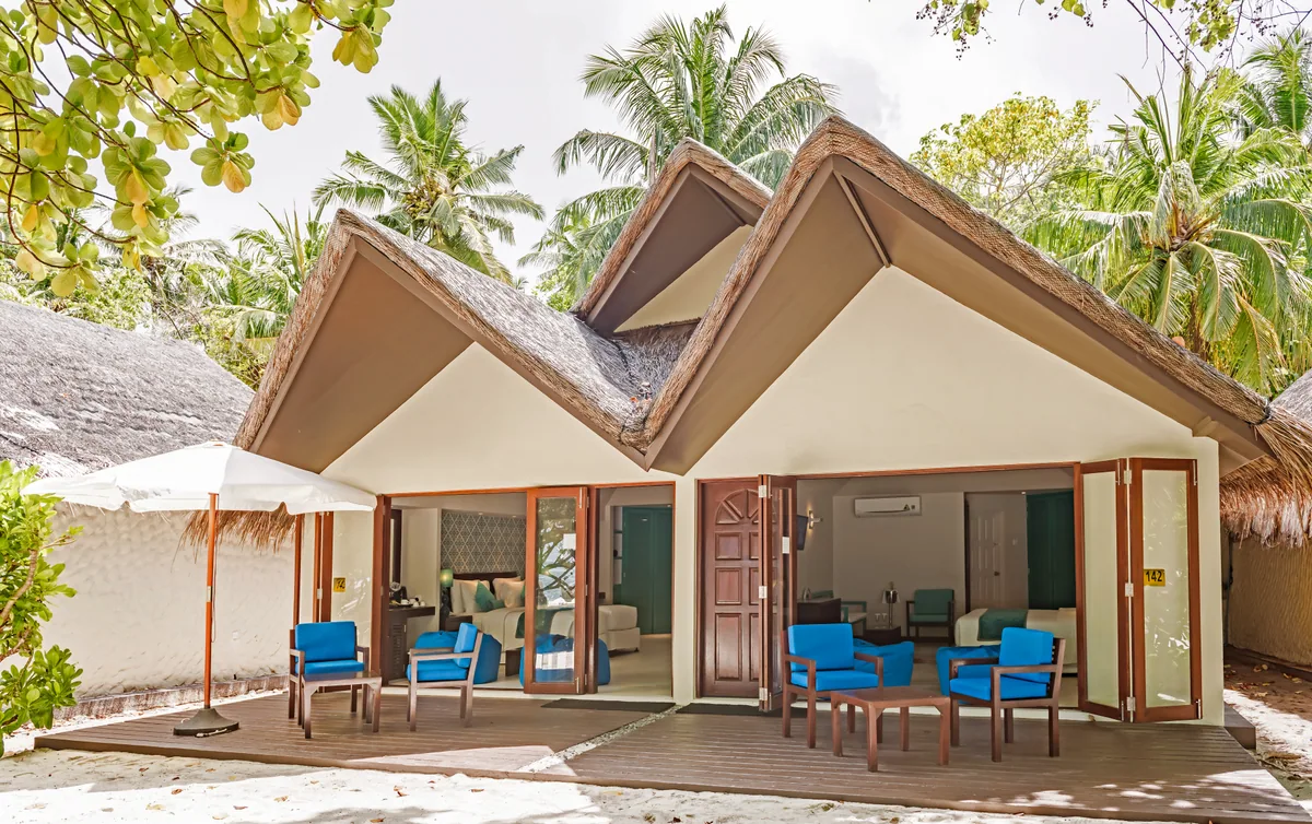 Adaaran Select Hudhuranfushi, villa beach, North Malé Atoll, Maldives, One-Bedroom Family Beach Villa exterior: a twin connected configuration with two identical thatched-pyramid-roof villas side-by-side, white walls with timber accents, dual entry points each with blue rattan armchairs on timber-deck verandas, white parasol at the left, sandy garden setting under dense palm canopy.
