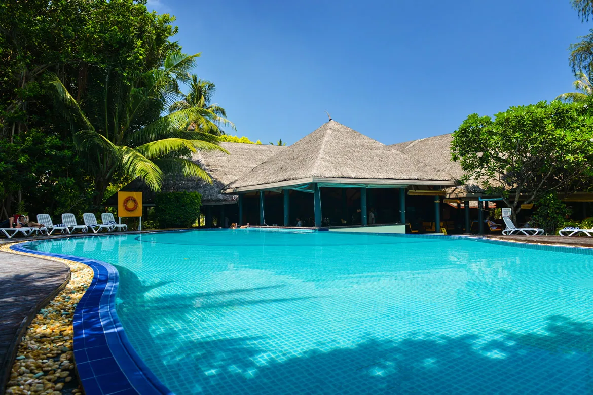Adaaran Select Hudhuranfushi, pool, North Malé Atoll, Maldives, Main pool: free-form pool with blue-mosaic-tile water surface set among the dense palm canopy, a thatched-roof poolside bar pavilion at the back edge, white sun loungers along the right side, a pebble border lining the pool edge, the all-inclusive resort's central recreation surface.