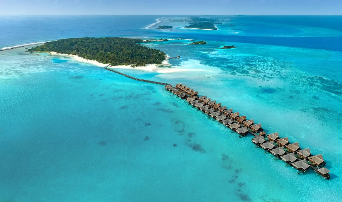 Adaaran Select Hudhuranfushi, aerial, North Malé Atoll, Maldives, Full property aerial: the long palm-covered Hudhuranfushi island stretching across the frame, the over-water villa row extending in a long curved arc into the lagoon (40 ocean villas configuration), the connecting walkway from the island, sand-spit beach at the bottom edge, neighbouring islands in the background, vast turquoise lagoon.