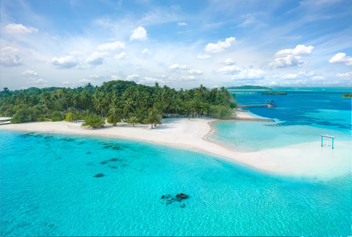 Adaaran Select Hudhuranfushi, aerial, North Malé Atoll, Maldives, Beach sand-tip aerial: the dense palm canopy of the main island with a long white sand-tip beach extending into the bright turquoise lagoon, a single over-water swing visible at the right edge (signature property lifestyle feature), a coral patch in the foreground water, the OWV jetty plus dive pavilion glimpsed at the right edge.