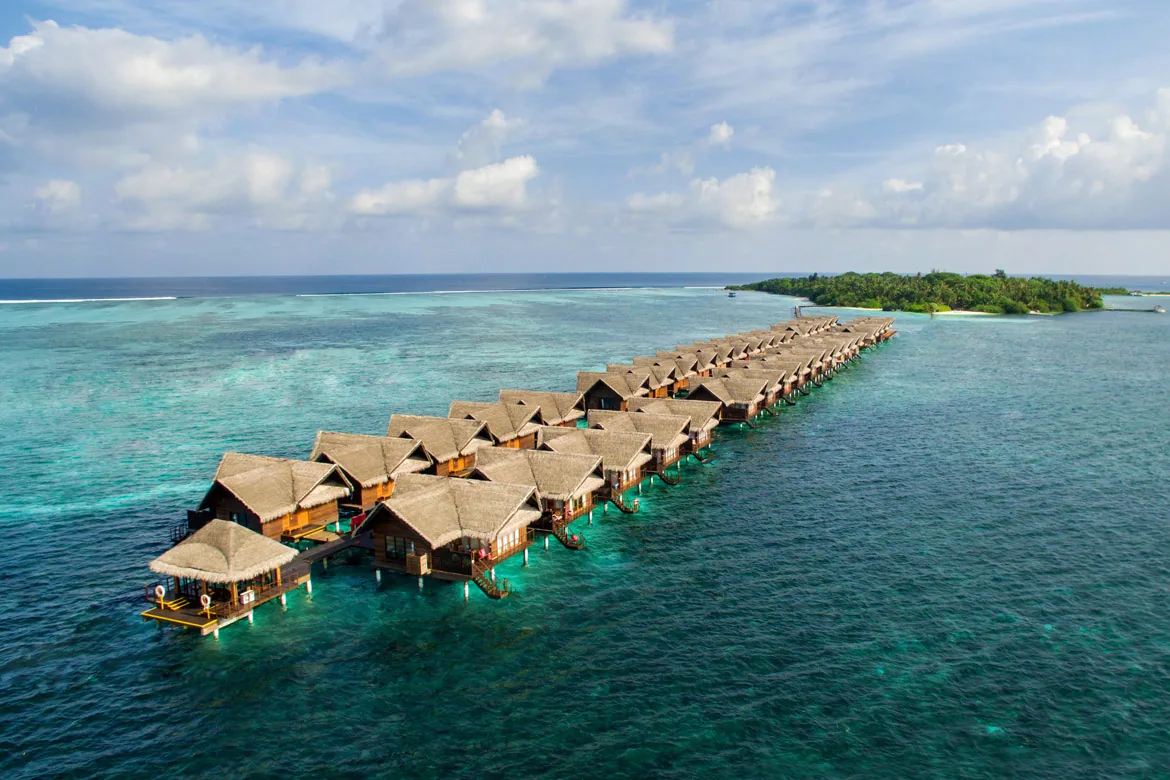 Adaaran Select Hudhuranfushi, villa overwater, North Malé Atoll, Maldives, Ocean Villa row low-altitude aerial: the full Ocean Villa configuration with thatched-roof villas in a long curved double-row formation on stilts over the lagoon, the connecting walkway extending from the palm-covered island visible at right, deep blue ocean to the left transitioning to turquoise reef visible beneath the villas.
