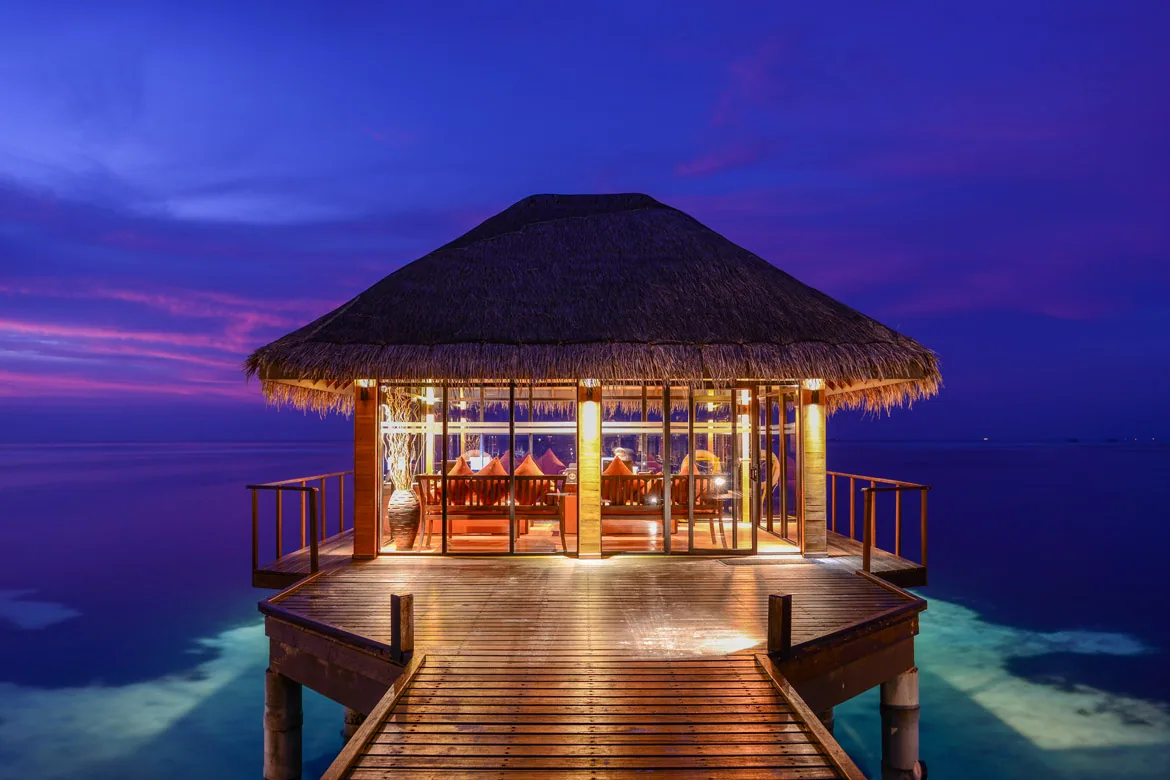 Adaaran Select Hudhuranfushi, dining, North Malé Atoll, Maldives, Over-water à-la-carte restaurant at twilight: thatched-roof pavilion at the end of a timber jetty, fully glass-walled with the warm-lit dining interior visible (orange seating, table lamps glowing through the glass), deep purple twilight sky reflecting on the perfectly calm lagoon, the speciality dinner-only venue captured in dusk framing.