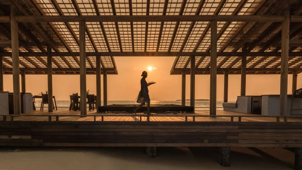 Alila Kothaifaru Maldives, The Shack sandbank sunset with lattice-roof pavilion, server silhouette carrying tray, central daybed and golden-hour ocean horizon, Raa Atoll, Maldives