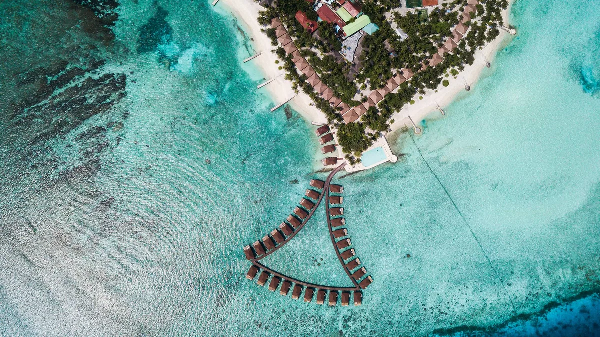 Alimathaa Maldives Resort, aerial, Vaavu Atoll, Maldives, Top-down aerial of the V-shape over-water villa cluster meeting at the wooded island tip in Vaavu Atoll, signature red-tile pyramid roofs lined along the curved boardwalk, reef edge at left.