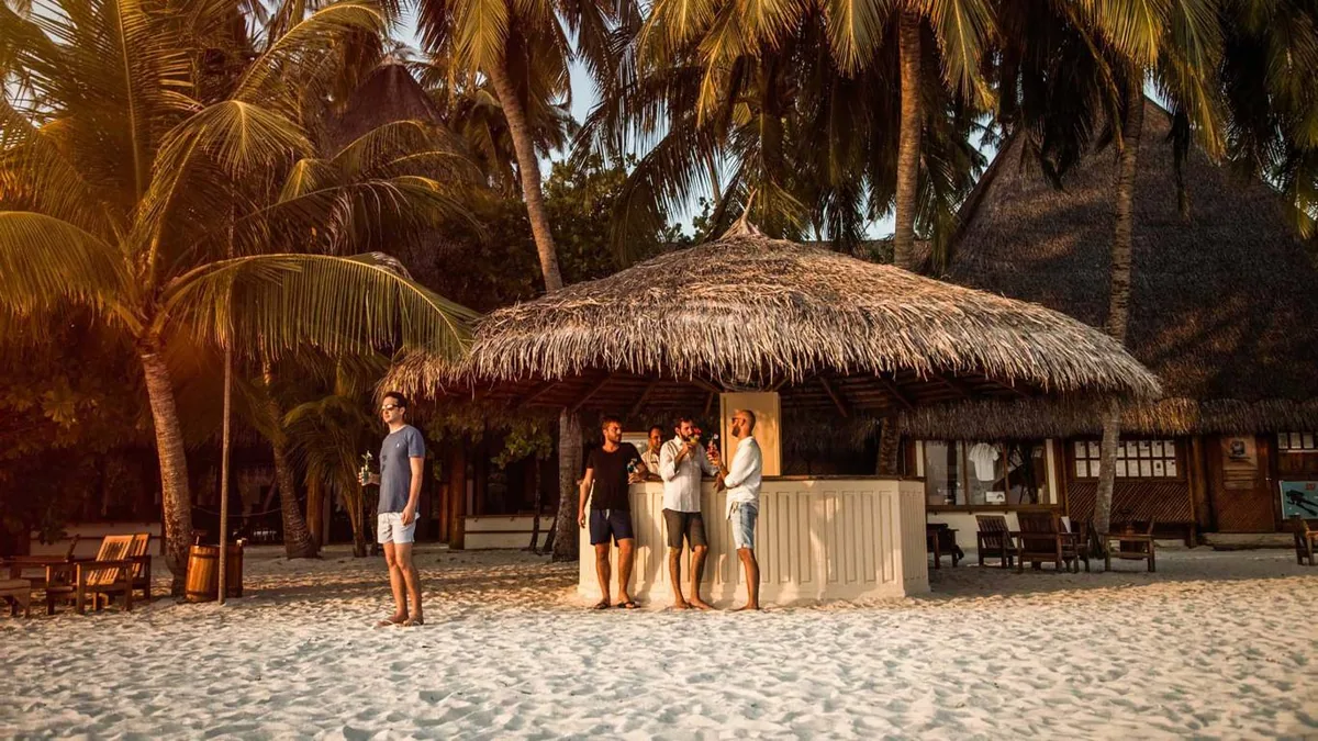 Alimathaa Maldives Resort, dining, Vaavu Atoll, Maldives, Beach bar pavilion at golden hour, round thatched-roof bar structure with guests gathered at the counter, palm-canopy backdrop and white sand around the perimeter.