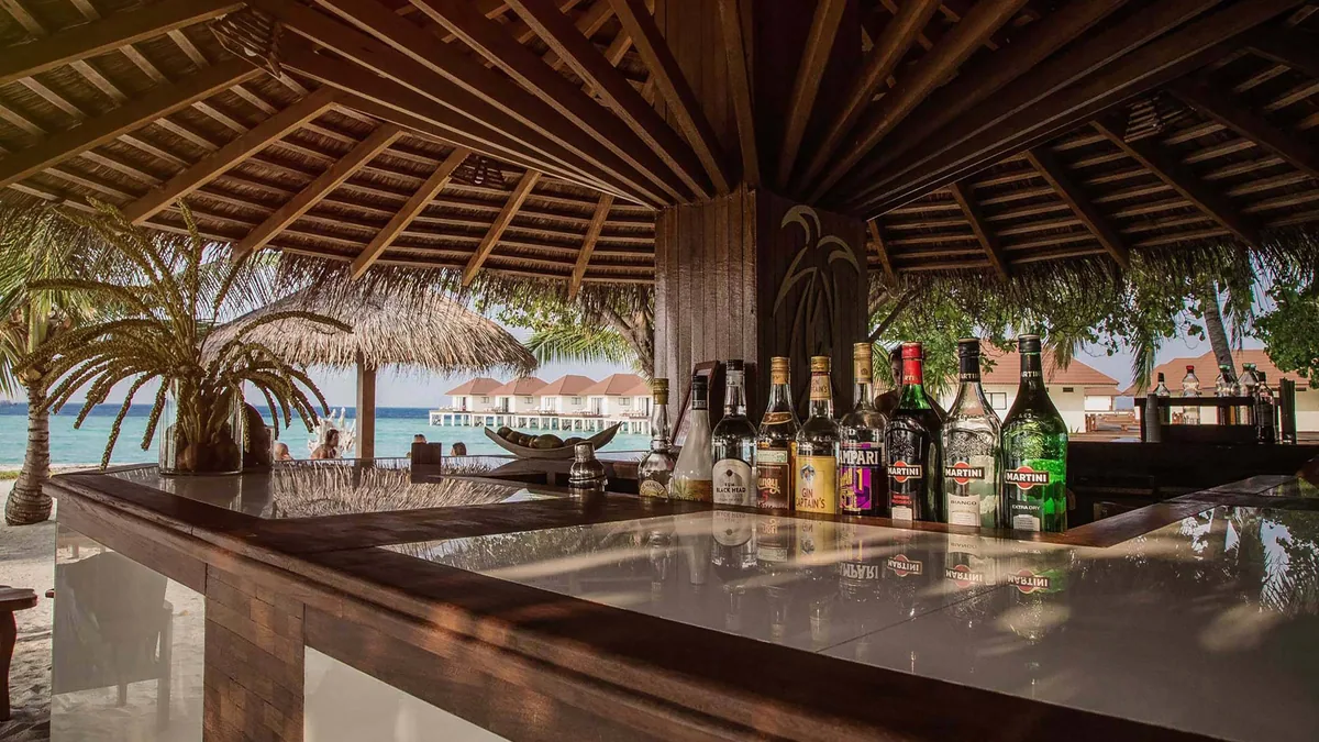 Alimathaa Maldives Resort, dining, Vaavu Atoll, Maldives, Bar counter close-up, Italian aperitivo bottles (Martini, Campari) lined along the timber counter, thatched ceiling overhead, view through to the over-water villa pavilions on the lagoon beyond (the property's Italian aperitivo hour, visible at the bar).