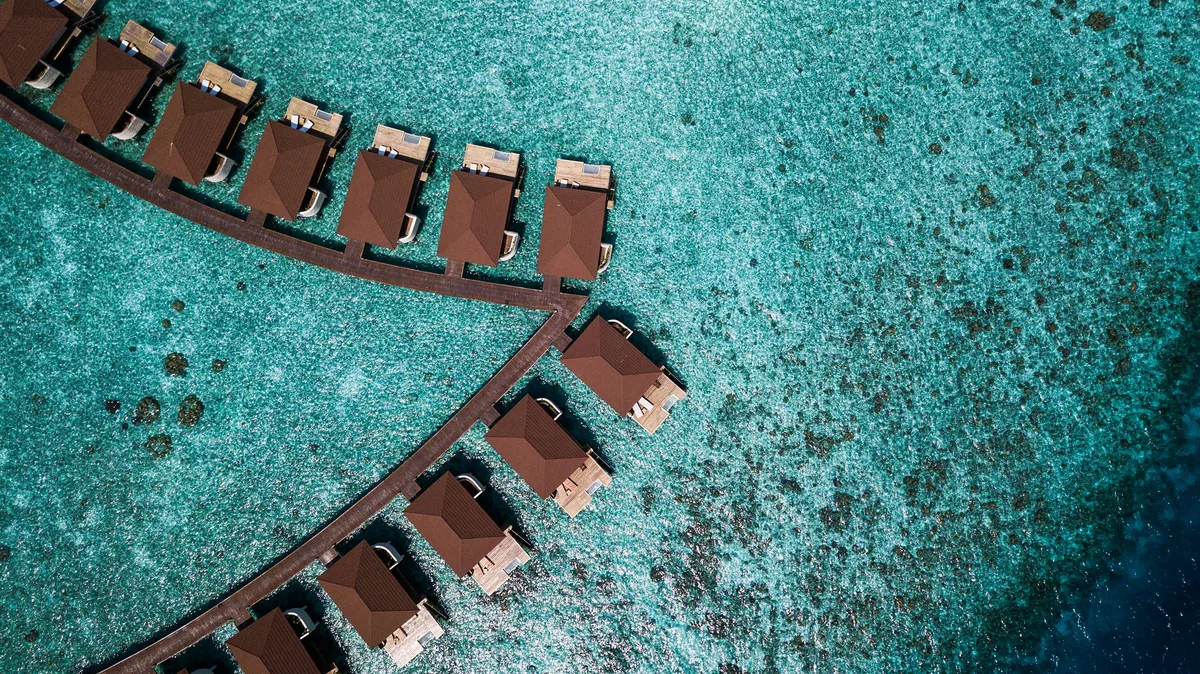 Alimathaa Maldives Resort, aerial, Vaavu Atoll, Maldives, Top-down close-up of two arched rows of over-water villas with their red-tile pyramid roofs aligned along the connecting walkway over a turquoise reef-flat.
