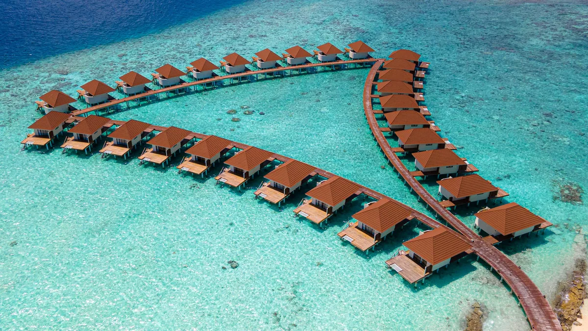Alimathaa Maldives Resort, aerial, Vaavu Atoll, Maldives, Oblique aerial perspective of the full V-shape over-water villa cluster with the timber boardwalk forming the apex, red-tile pyramid roofs lined symmetrically along each arm above the lagoon.