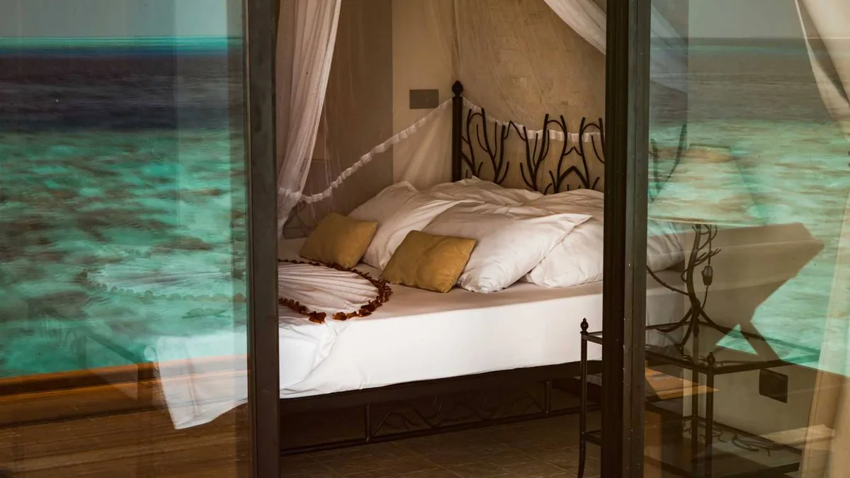Alimathaa Maldives Resort, villa interior, Vaavu Atoll, Maldives, Over-water villa bedroom, a wrought-iron coral-motif four-poster bed (the property's signature interior detail) with white linens and yellow throw pillows, mosquito-net canopy, rose-petal heart on the bed, reef visible through glass doors on both sides.