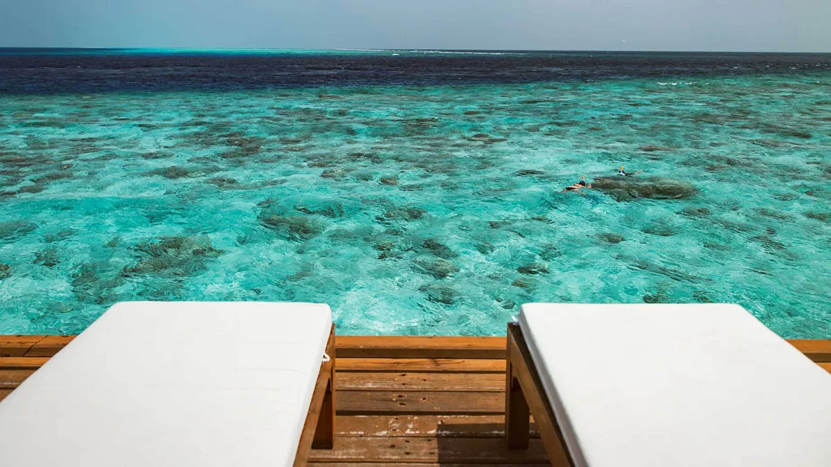 Alimathaa Maldives Resort, setting, Vaavu Atoll, Maldives, Reef view from the over-water villa deck, a snorkeler in the clear water above the coral garden, sun loungers on the timber deck at the edge of the frame, lagoon-edge reef stretching to the channel beyond.
