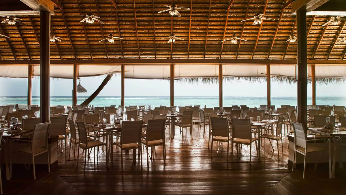 Alimathaa Maldives Resort, dining, Vaavu Atoll, Maldives, Principal pavilion interior, wide open-sided thatched A-frame with ceiling fans, mesh-and-aluminium chairs around dining tables on polished timber floor, the lagoon visible through the seaward open frame.