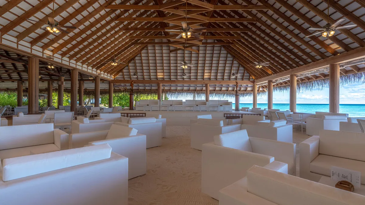 Alimathaa Maldives Resort, dining, Vaavu Atoll, Maldives, Open-air bar lounge pavilion, exposed timber A-frame ceiling with ceiling fans, white sofa clusters arranged on a sand floor (the property's signature lounge detail), thatched palm-fringe perimeter opening to the lagoon at right.