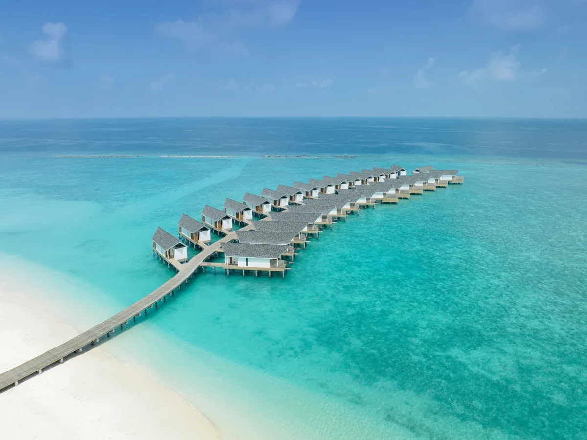 RAAYA by Atmosphere, double-row over-water villa cluster with pitched shingle-tile roofs along jetty from sand peninsula on turquoise lagoon, Raa Atoll, Maldives