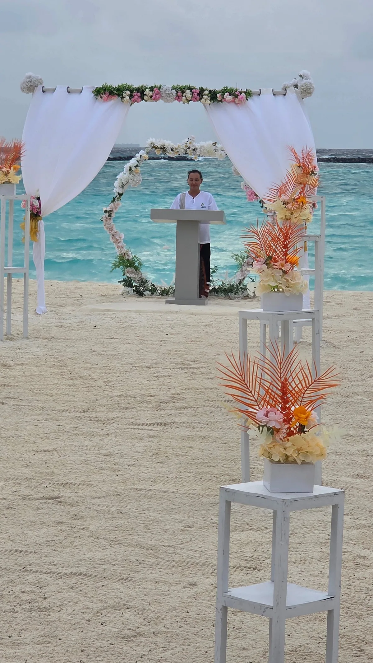 RAAYA by Atmosphere, beach wedding ceremony setup with white-draped circular arch, floral garland, officiant lectern and orange tropical flowers in pedestal vases, Raa Atoll, Maldives