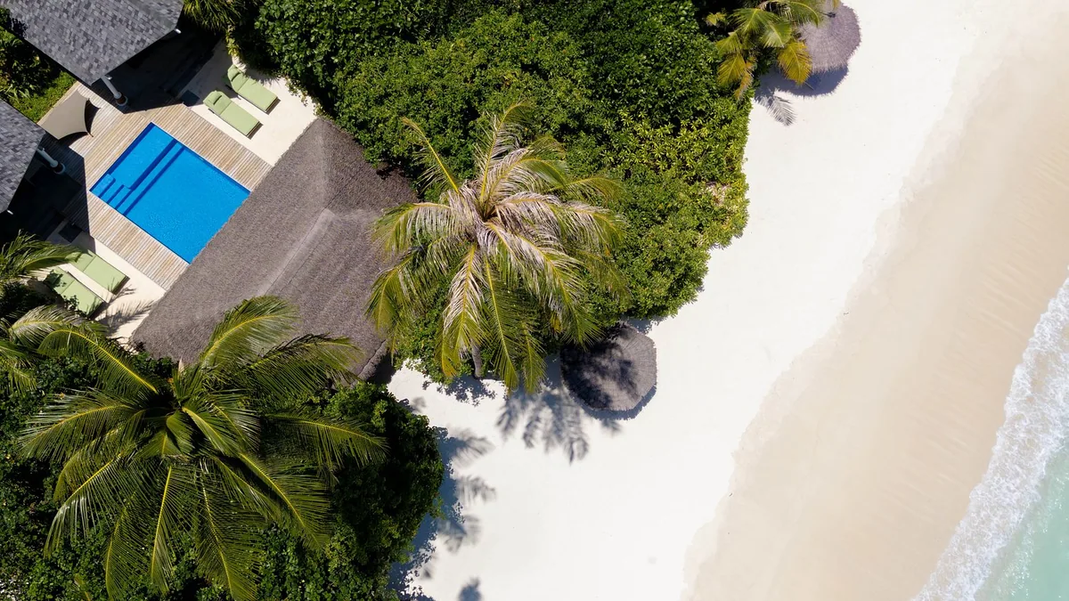 RAAYA by Atmosphere, Two Bedroom Family Beach Villa with Pool top-down aerial showing thatched roof, rectangular blue pool, four green sun loungers on palm-shaded deck, Raa Atoll, Maldives