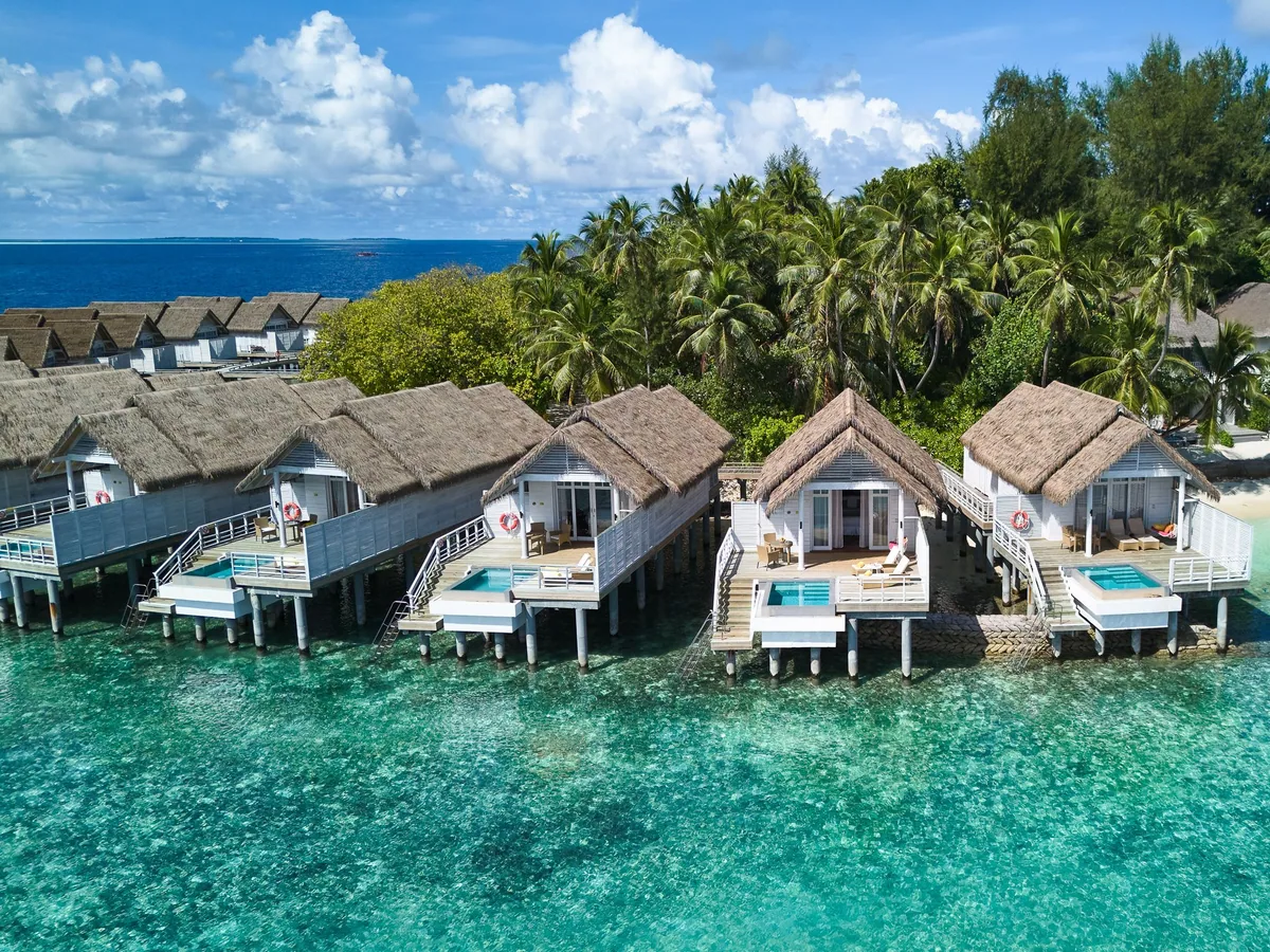 Amaya Kuda Rah Maldives, villa overwater, Alif Dhaal (South Ari) Atoll, Maldives, Overwater villa row aerial close-up, the thatched-roof villas with white walls extending into the lagoon along the boardwalk, deck steps descending into the clear water, palm canopy of the main island visible behind.
