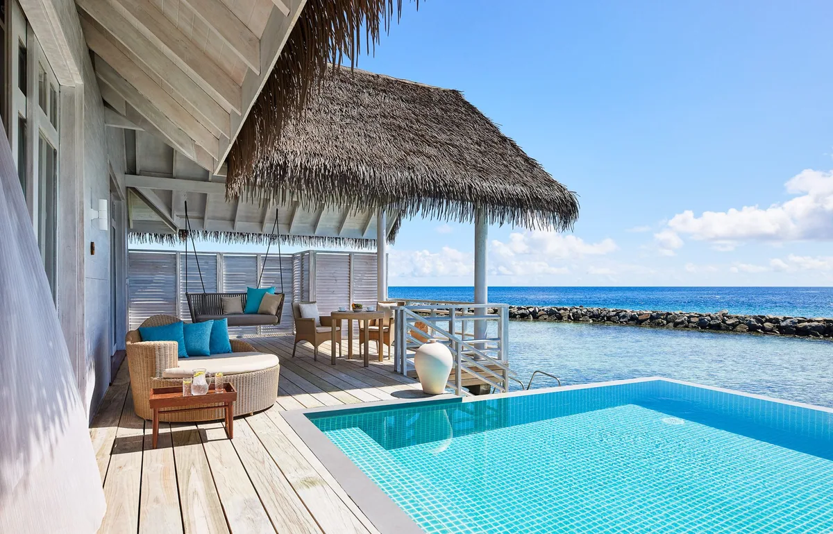 Amaya Kuda Rah Maldives, villa overwater, Alif Dhaal (South Ari) Atoll, Maldives, Over Water Suite with Private Pool deck, thatched eaves overhead with hanging-rope swing chair and wicker daybed loungers, dining table-and-chairs, blue mosaic-tile infinity-edge pool stretching toward the lagoon and ocean horizon, reef-protection wall visible.