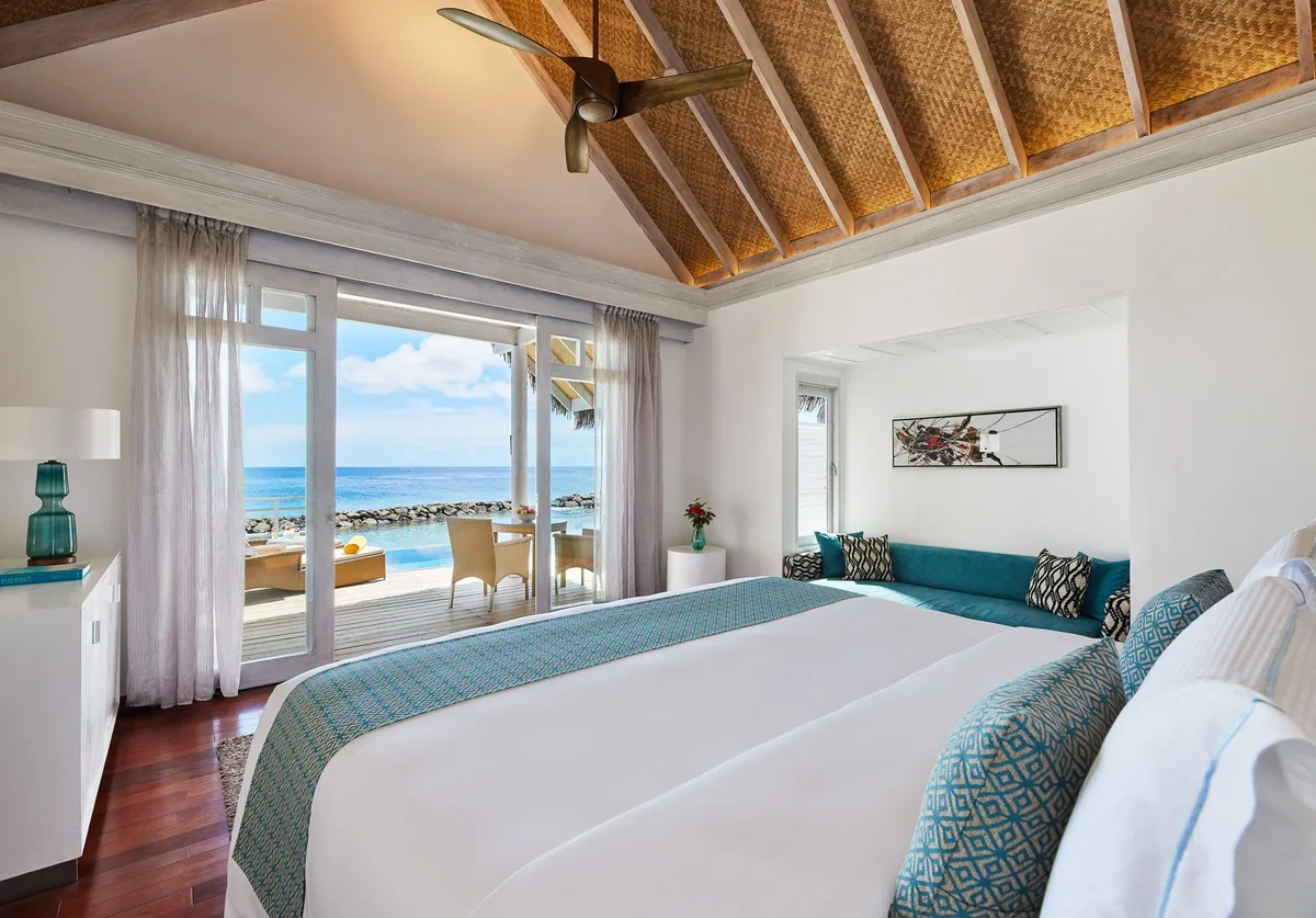 Amaya Kuda Rah Maldives, villa overwater, Alif Dhaal (South Ari) Atoll, Maldives, Overwater Villa interior, vaulted woven-bamboo ceiling with timber rafters and ceiling fan, king bed with teal accent throw and matching square cushions on the banquette, retractable glass doors opening to the deck with sun-loungers, dining table and the sea horizon beyond.