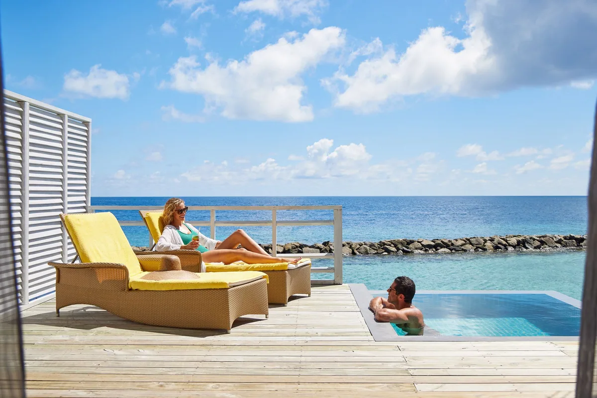 Amaya Kuda Rah Maldives, lifestyle, Alif Dhaal (South Ari) Atoll, Maldives, Couple on the overwater villa deck, woman in white reading on the yellow-cushioned woven sun-lounger while the man cools off in the infinity-edge plunge pool, woven-slat privacy panel framing the deck, reef-protection wall and deep blue horizon beyond.