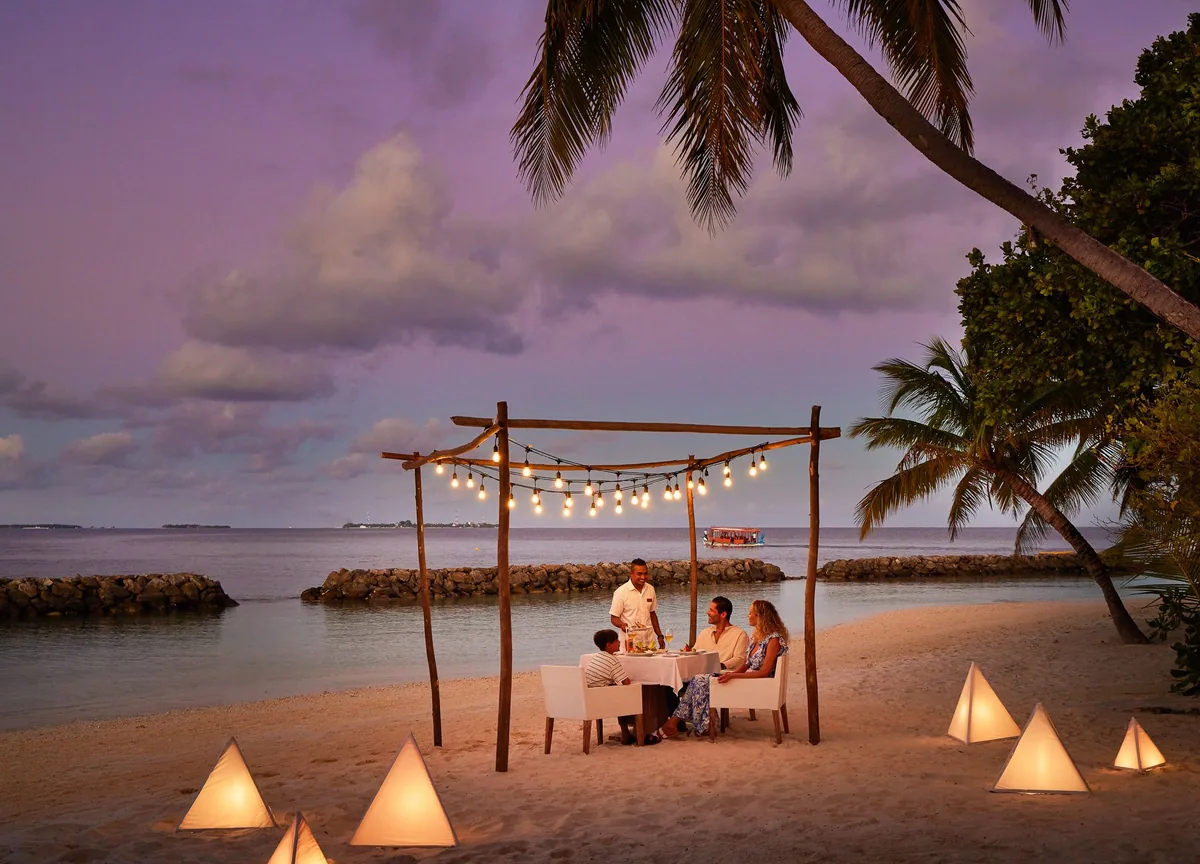 Amaya Kuda Rah Maldives, dining, Alif Dhaal (South Ari) Atoll, Maldives, Family private beach dining at twilight, family of four seated at the table under a wooden-frame fairy-light canopy, a butler serving from the side, pyramid paper-lantern candles arranged on the white sand, palm fronds framing and the purple-pink dusk sky over a distant atoll silhouette.