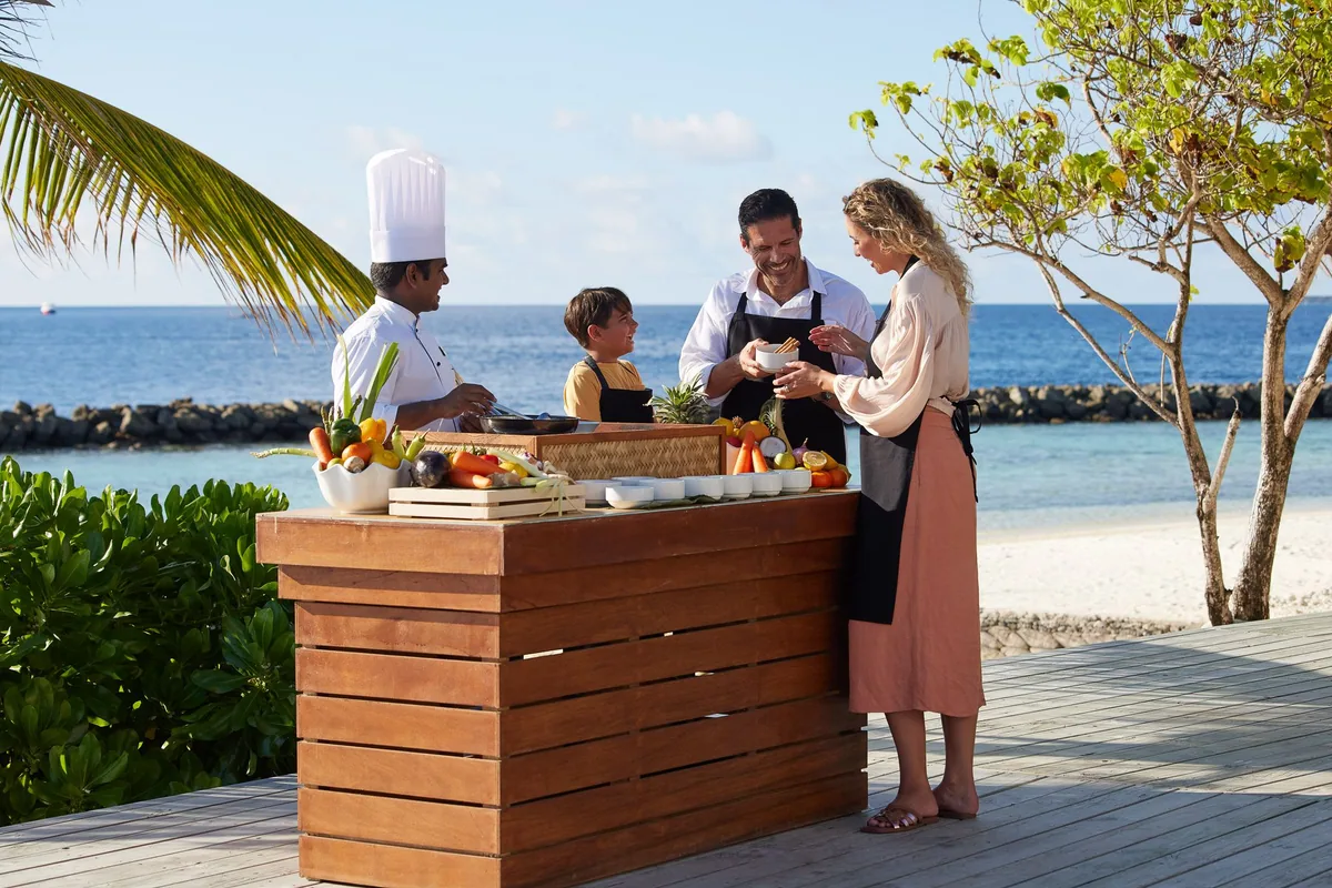 Amaya Kuda Rah Maldives, activities, Alif Dhaal (South Ari) Atoll, Maldives, Cooking class on the beach deck, chef in white toque with a family of three at the outdoor counter handling vegetables and tropical fruit, palm fronds and the lagoon edge framing. The Aitken Spence Sri-Lankan-cuisine integration at the experiential layer.