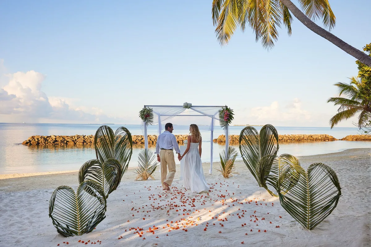 Amaya Kuda Rah Maldives, lifestyle, Alif Dhaal (South Ari) Atoll, Maldives, Beach wedding altar, bride-and-groom approaching the chuppah-style flower-decorated arch on the white sand, dual palm-frond heart sculptures flanking the rose-petal-strewn aisle, reef-protection wall and deep blue lagoon framing the horizon. The property's romance-and-weddings programme as one of the booking pillars.