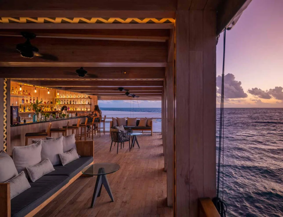 Amilla Maldives, dining, Baa Atoll, Maldives, Sunset Bar wood-clad interior with the full bottle wall behind the curved counter, the yellow-zigzag fascia along the wood-beam ceiling, cushion-banquette lounge alcove, and the open wall framing the dusk horizon with rope-rigging detail.