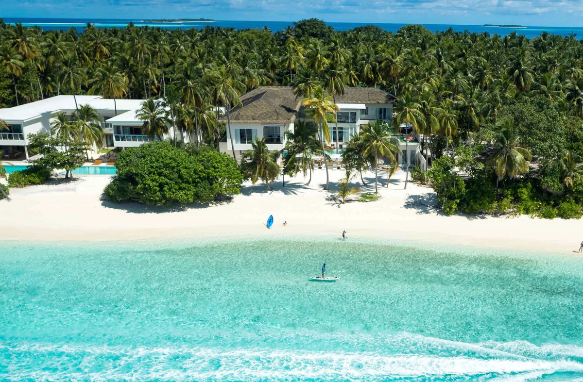 Amilla Maldives, villa beach, Baa Atoll, Maldives, Beach-front residence aerial with the multi-storey white architecture and pyramid-roof centre block, the broad palm-canopy backdrop, the white-sand beach, and two SUP paddlers in the foreground turquoise lagoon.