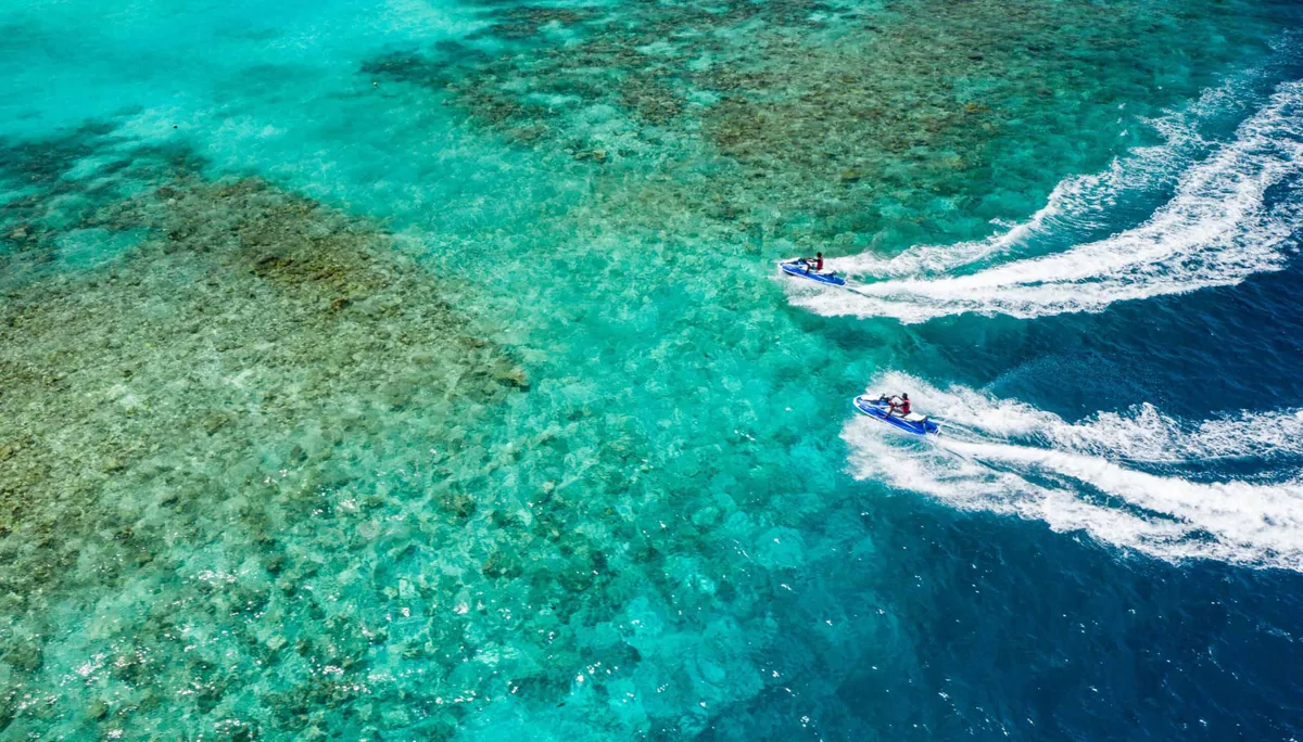 Amilla Maldives, activities, Baa Atoll, Maldives, Two jet skis carving figure-eight wake patterns across the deep-blue lagoon shelf with the vivid reef shadow patterns visible beneath translucent turquoise water at the top-down POV.