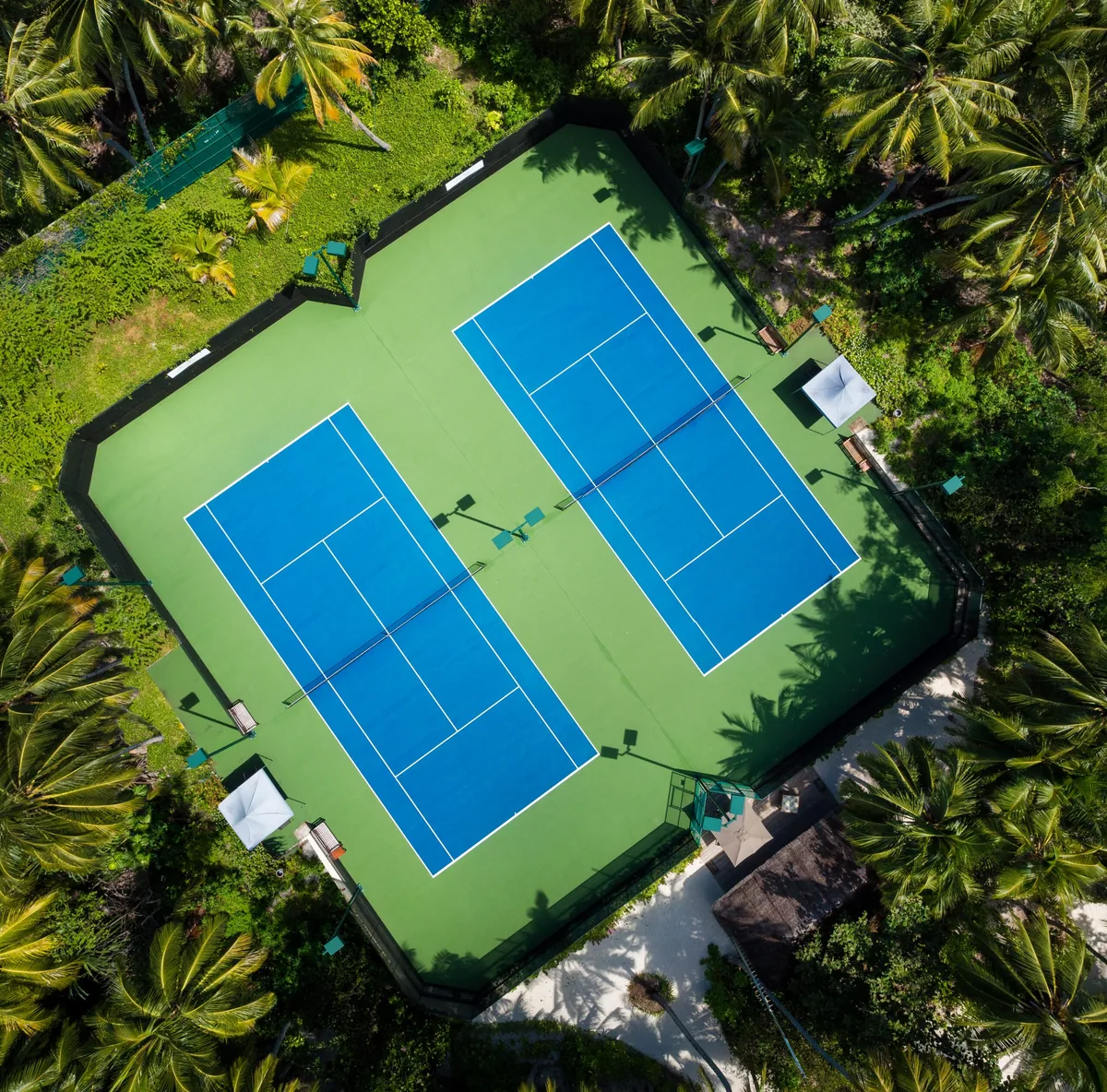 Amilla Maldives, activities, Baa Atoll, Maldives, Twin tennis courts aerial with the blue-on-green hard surface embedded in the dense palm canopy on Amilla Fushi island, the property-distinctive amenity rare among Maldivian resorts.