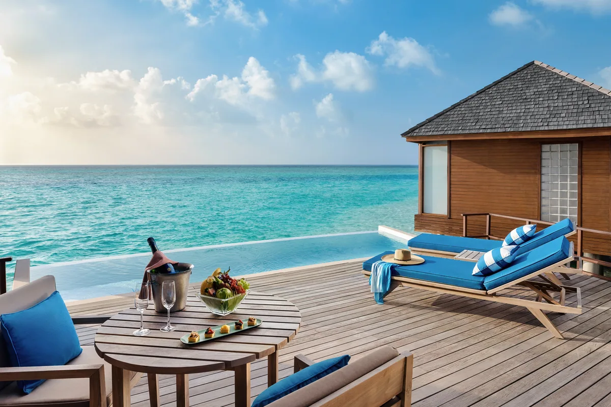 Anantara Dhigu Maldives Resort, villa overwater, South Malé Atoll, Maldives, Deluxe Sunset Over-Water Pool Villa deck, the private infinity-edge plunge pool merging into the open turquoise lagoon, twin blue-cushioned teak loungers facing the ocean, a champagne-and-canapés tray on the lounge table, blue-and-white striped cushions, and the wood-shingle villa wall at the right.