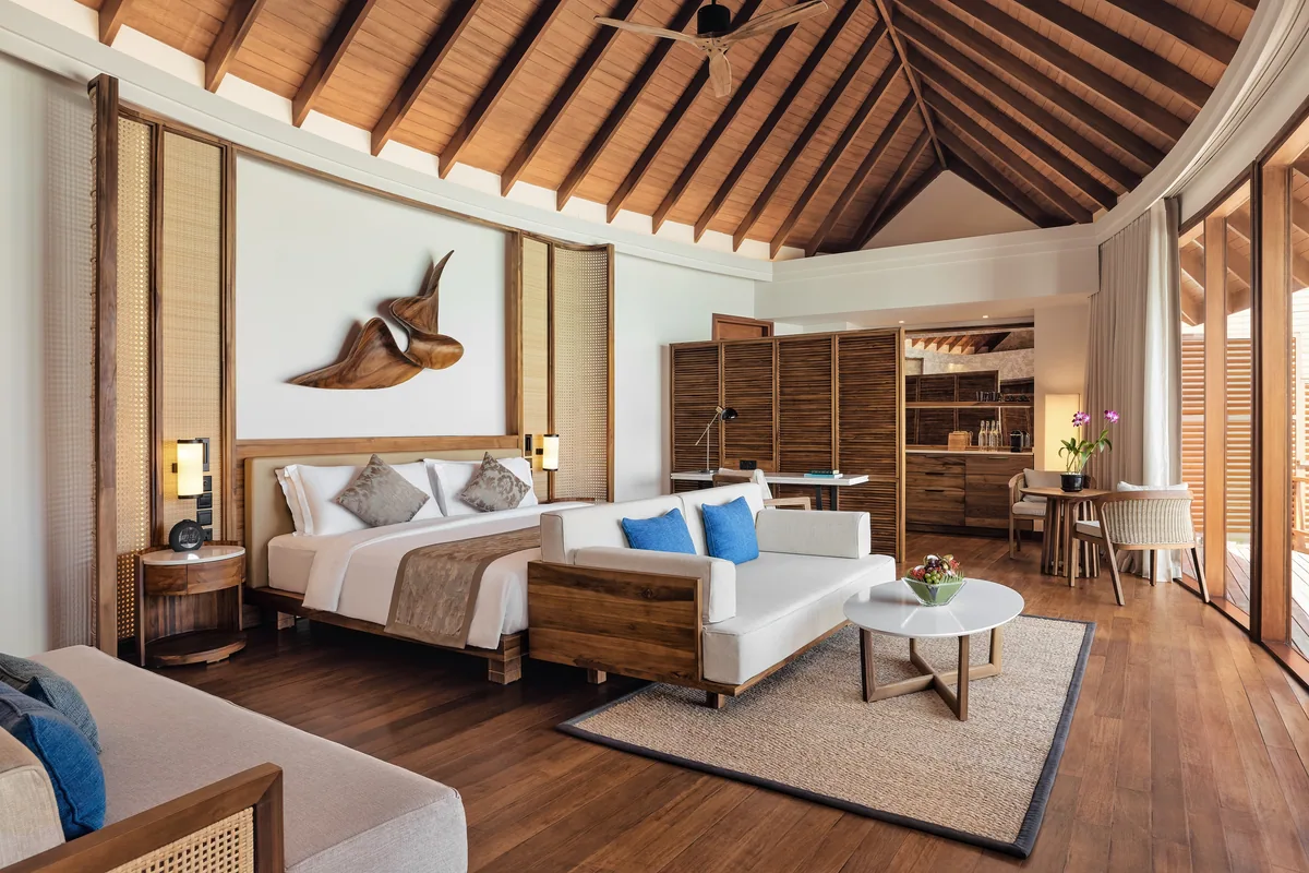 Anantara Dhigu Maldives Resort, villa overwater, South Malé Atoll, Maldives, Renovated Over-Water Villa bedroom, the vaulted hardwood-rafter ceiling, the signature carved-wood MANTA RAY wall sculpture above the bed (the Anantara Dhigu signature interior motif), woven-cane screen panels flanking the bed, contemporary Maldivian-modern teak furniture, hardwood floor, and the open-deck dining area visible through the corner glazing.
